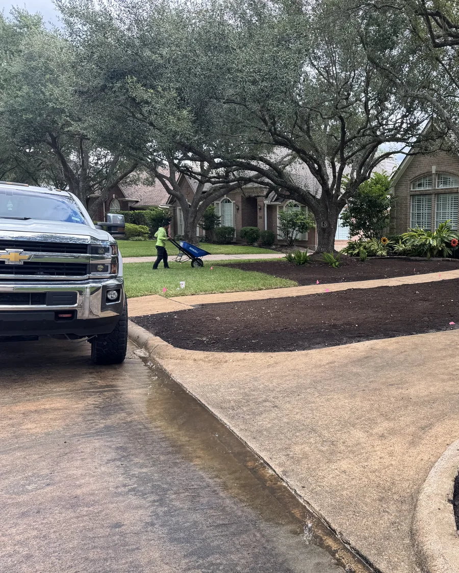 A crew member moving a wheelbarrow across the lawn while fresh beds are mulched along the driveway